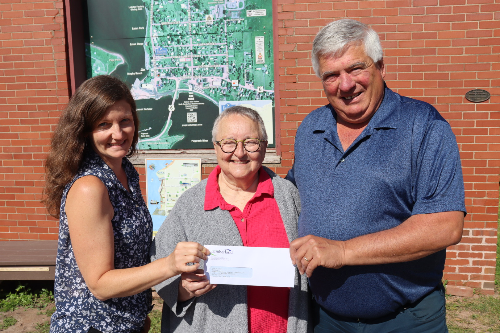 Two women and a man stand together holding a cheque in front of brick building.